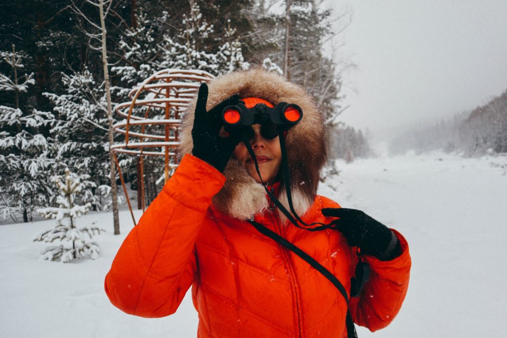 a woman in an orange jacket in the snow looking through a pair of binoculars