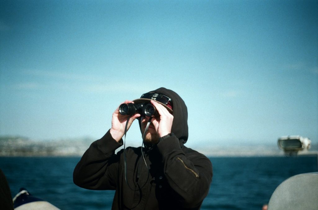 A man wearing a black hoodie using binoculars on a boat