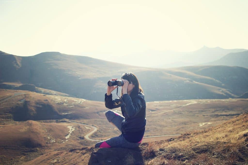 A woman wearing black jacket and jogging pants sitting on a rock and using binoculars