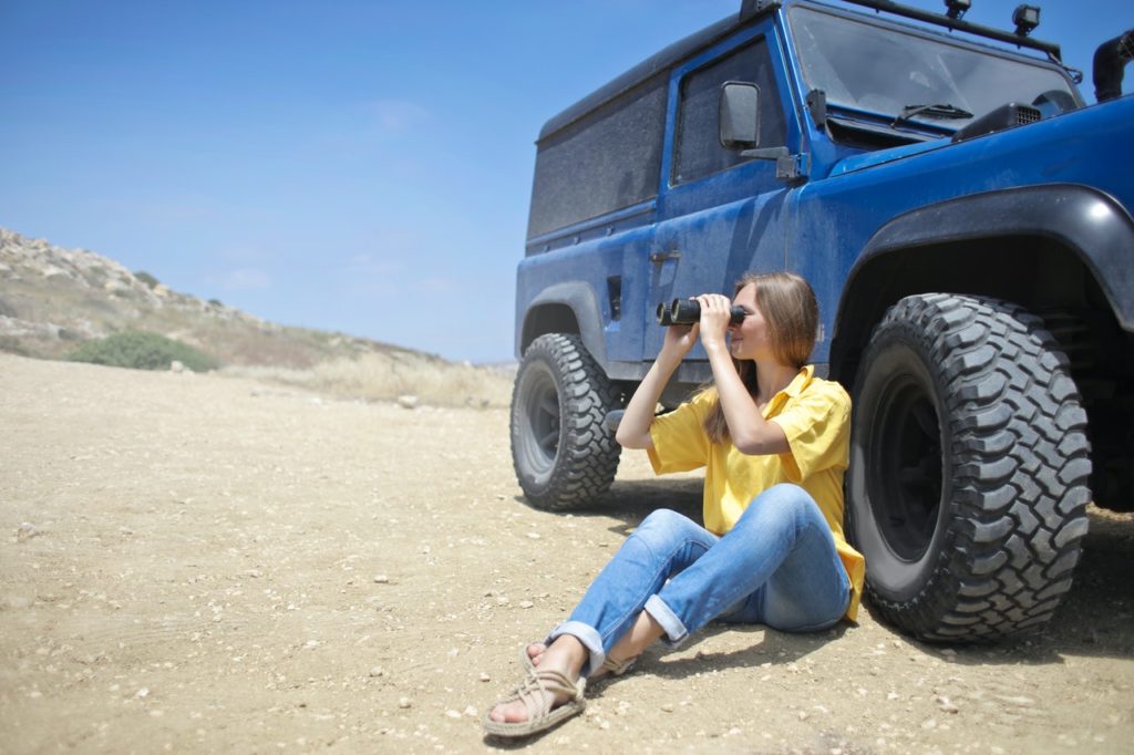 W woman sitting on the ground next to a blue jeep and looking through a pair of binoculars